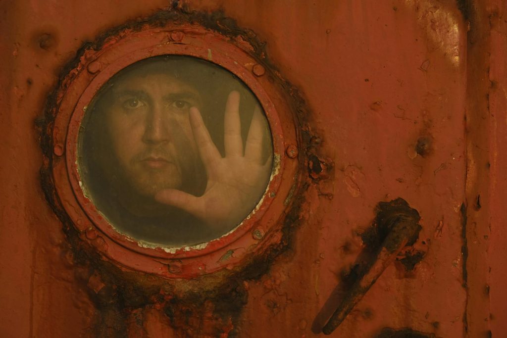 A man peers through a round, rusty porthole window, creating a mysterious, eerie atmosphere.
